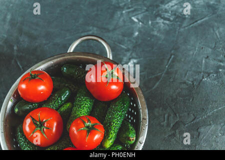 Rohe ganze gewaschene Tomaten und Gurken in einem Sieb auf einem dunklen konkreten Hintergrund. Blick von oben. Flach. Stockfoto