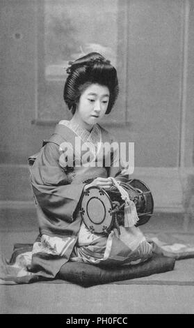 [1920s Japan - Geisha mit einer Tsutsumi Drum] - die Geisha im Kimono und traditionelle Frisur hält ein tsutsumi (auch Tsuzumi) Schulter drum. Die gleiche Art der Fässer wurden in Noh und Kabuki Dramen verwendet. Die tsutsumi wird mit der linken Hand auf der rechten Schulter gehalten, während die rechte Hand verwendet wird. Das Gerät verfügt über zwei Felle durch Spannung Kabel unterstützt. Diese Kabel sind gezogen, um die Tonhöhe zu ändern. 20. jahrhundert alte Ansichtskarte. Stockfoto