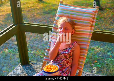 Das kleine Mädchen sitzt auf einem Liegestuhl auf einer Veranda. Kleine Mädchen ist Essen Käse aromatisiert Snacks im Garten. Verträumt und romantisch Bild. Sommer und glückliche Kindheit Konzept Stockfoto