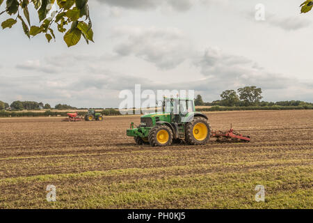 Großbritannien Landwirtschaft, zwei Traktoren von John Deere, die bei der Arbeit Tiefenlockerung und Drillen einem Abgeernteten Feld Stockfoto