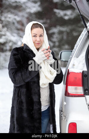 Europäische Frau mit warme Kleidung friert im Winter Holz in der Nähe von Broken Auto, trinken heißen Getränk Stockfoto