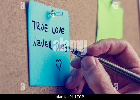 Nahaufnahme der männlichen Hand schreiben Wahre Liebe stirbt nie die Phrase auf blauen Post it Papier auf Kork Pinnwand, retro Effekt blass aussehen. Stockfoto