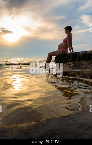 Schwangere Frau tragen Bikini am Ufer auf den Felsen sitzen Zurückgelehnt genießen einen schönen Sonnenuntergang. Stockfoto