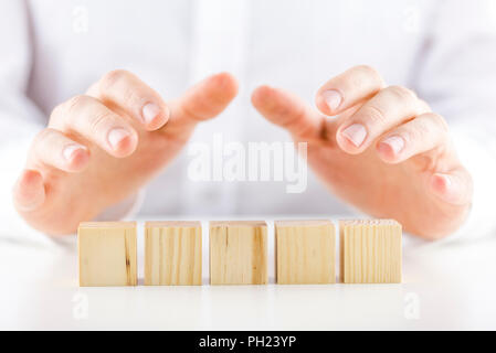 Mann hält seine Hand schützend über eine Reihe von fünf leeren Holzwürfel auf einem weißen reflektierenden Tabelle in einem konzeptionellen Bild. Bereit für Ihren Text. Stockfoto