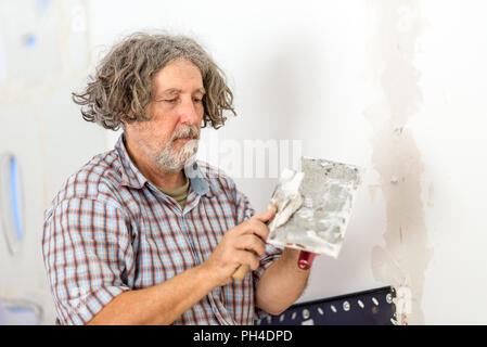 Im mittleren Alter männlich Bauherr oder Hausbesitzer die Reparatur einer Anwendung Füller Putz von der Wand eine kleine Platine mit einem Schaber, als er die Reparatur eines Risses oder öffnen. Stockfoto