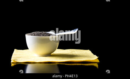 Schüssel mit gesunden Chia Samen, die reich an Omega-3-Fettsäuren mit einem Löffel auf einer schwarzen reflektierende Tisch vor einem schwarzen Hintergrund mit Copyspace. Stockfoto