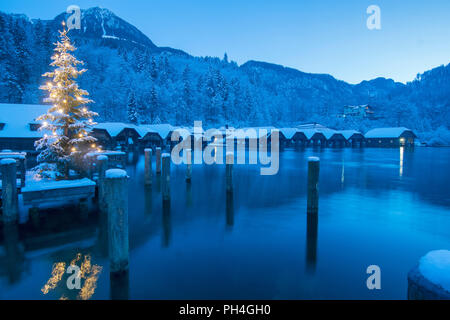 Die leuchtenden Weihnachtsbaum am Seelaende am See Königssee mit Boot Hütten im Hintergrund. Berchtesgadener Land, Bayern, Deutschland Stockfoto