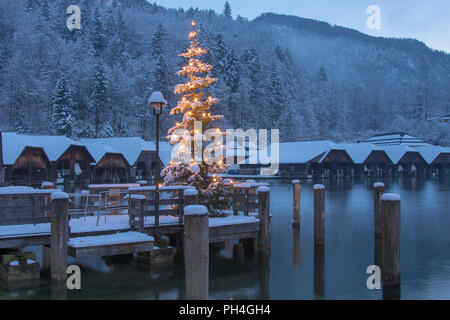 Die leuchtenden Weihnachtsbaum am Seelaende am See Königssee mit Boot Hütten im Hintergrund. Berchtesgadener Land, Bayern, Deutschland Stockfoto