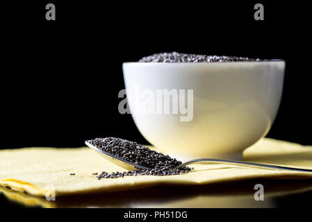 Aromatische gesunde Chia Samen von Salvia hispanica Pflanze reich an Omega-3-Fettsäuren in einem Löffel neben einem weißen Keramik Schüssel gefüllt mit Chia auf Stockfoto