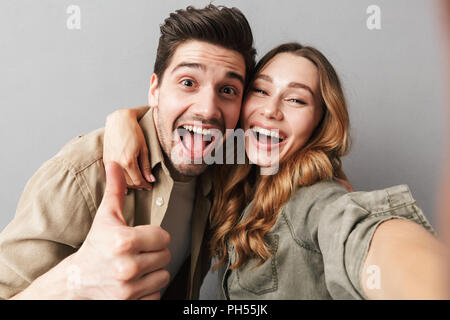 Portrait einer aufgeregt, junges Paar umarmen sich während einer selfie und Daumen hoch über grauer Hintergrund Stockfoto