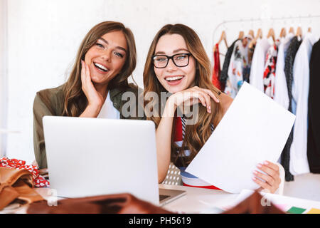 Zwei freudige junge Frauen Kleidung Designer zusammen arbeiten im Atelier, am Tisch mit Laptop und Entwürfe sitzen Stockfoto
