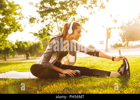 Bild von lächelnden Sportlerin 20 s Kopfhörer arbeiten und Strecken die Beine beim Sitzen auf der Trainingsmatte im Green Park Stockfoto