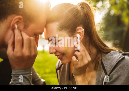 Bild der glücklichen jungen Menschen Mann und Frau 20 s in Trainingsanzügen hören zusammen Musik über Kopfhörer beim Gehen im Green Park Stockfoto