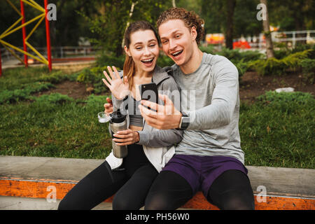 Foto fröhliche, sportliche Paar Mann und Frau 20 s in Sportkleidung sitzen auf Attika im Green Park und unter selfie nach dem Workout oder Jog an sonnigen Summe Stockfoto