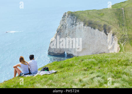 Ein paar setzte sich auf einem grasbewachsenen Steilküste auf einem sonnigen klaren Tag, bis zu einer Bucht von Fledermäusen den Kopf, in der Nähe der Durdle Door auf der Jurassic Coast South England Stockfoto
