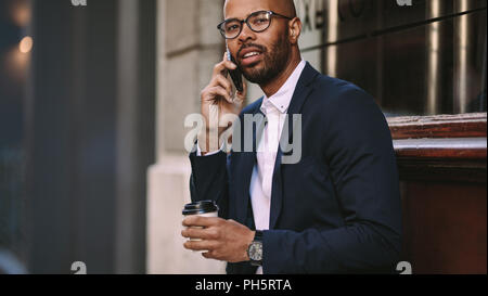 Portrait von hübscher junger Geschäftsmann einen Anruf beim Sitzen im Freien mit einem Kaffee. Mann in Anzug reden über Handy. Stockfoto