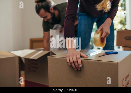 Paar mit Kisten packen, den Hausrat zu packen. Mann setzen Objekte in Kisten, während eine Frau Klebeband auf die Boxen gilt, um sie zu verschließen. Stockfoto