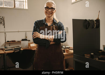 Portrait von zuversichtlich älteren weiblichen Juwelier stehen in Ihrer Werkstatt. Frau Goldsmith tragen Schürze und Brillen Stehen mit verschränkten Armen und Stockfoto