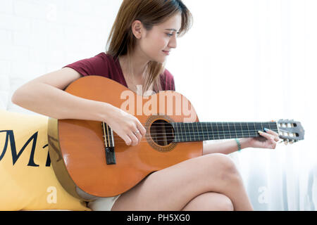 Junge schöne asiatische Frau sitzt auf einem Sofa, Gitarre zu spielen - Mädchen Musik, Spaß Konzept Stockfoto