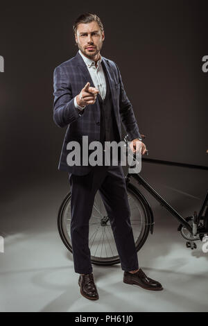 Ernster junger Mann in Anzug, der auf der Kamera beim Stehen in der Nähe von Fahrrad auf Schwarz Stockfoto