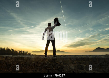 Geschäftsmann Holding aloft seine Jacke bei Sonnenuntergang, als er an einer Wand mit Blick auf einen Misty Mountain Szene steht Silhouette gegen den farbenfrohen Himmel w Stockfoto