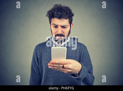 Junge stirnrunzelnd angewidert Mann surfen Smartphone in Abneigung auf grauem Hintergrund Stockfoto