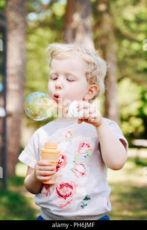 Portrait von niedlichen Lustige kleine blonde Kaukasier Kind girl Toddler, stehen im grünen Wald Feld Wiese bläst Seifenblasen, hellen Sommertag, su Stockfoto