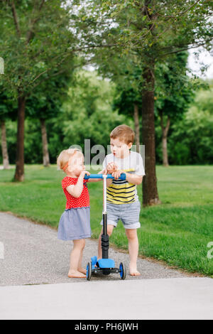 Zwei kleine Kaukasischen Vorschulkinder kämpfen im Park. Jungen und Mädchen können nicht ein scooter teilen. Ältere Geschwister Bruder nicht sein Spielzeug zu geben Stockfoto