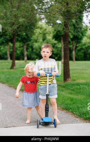 Zwei kleine Kaukasischen Vorschulkinder kämpfen im Park. Jungen und Mädchen können nicht ein scooter teilen. Ältere Geschwister Bruder nicht sein Spielzeug zu geben Stockfoto