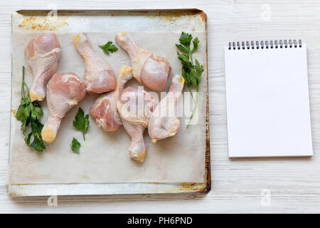 Ungekocht raw Hähnchenschenkel auf ein Backblech Papier in Fach und leere Notepad, Ansicht von oben. Weiß Holz- Hintergrund. Platz für Text. Stockfoto