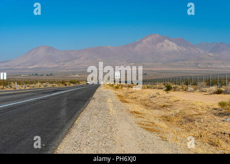 Wüste Autobahn mit Tempolimit Zeichen auf dem Weg zu hohen kahlen Berge führen unter einem Bight blauen Himmel. Kies am Straßenrand und tote Vegetation. Stockfoto