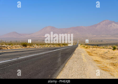 Wüste Autobahn mit Tempolimit Zeichen auf dem Weg zu hohen kahlen Berge führen unter einem Bight blauen Himmel. Kies am Straßenrand und tote Vegetation. Stockfoto