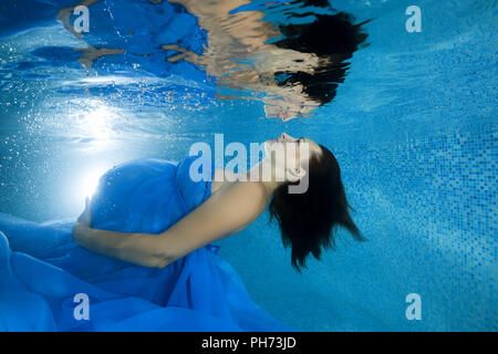 Eine schwangere Frau in einem blauen Kleid schaut sie Reflexion durch die Unterwasserwelt im Pool Stockfoto
