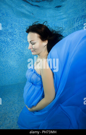 Schwangere Frau in einem blauen Kleid unter Wasser im Pool posiert Stockfoto