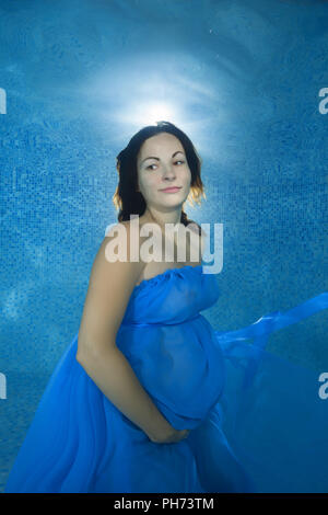 Schwangere Frau in einem blauen Kleid unter Wasser im Pool posiert Stockfoto