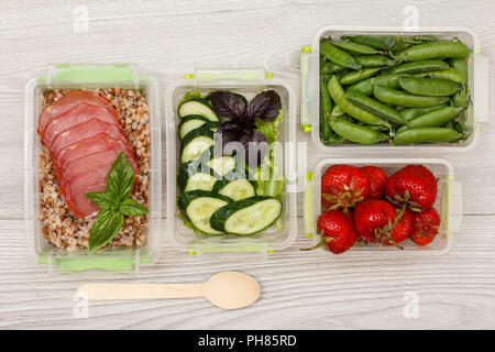 Kunststoff Mahlzeit prep Container mit gekochten Buchweizen Brei und Scheiben von Fleisch, frischen Gurken und Salat, Erdbeeren, grüne Erbsen und Löffel aus Holz o Stockfoto