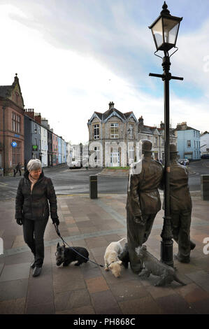 Eine Hundespaziererin lässt ihre Scottie-Hunde in der Laurel & Hardy-Statue Ulverston, Cumbria, dem Geburtsort von Stan Laurel im Jahr 1890, den beißenden Hund treffen Stockfoto