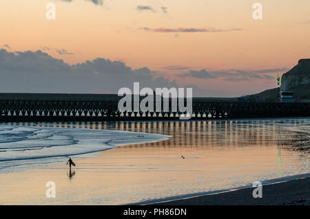 Nicht identifizierbare Person mit Surfbrett zu Fuß bei Ebbe auf Seaford Strand bei Sonnenuntergang, mit Reflexionen von Newhaven Hafen auf nassem Sand. Stockfoto