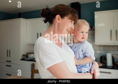 Seitenansicht einer Mutter, die ihre Tochter Holding. Sie sind beide schauen aus dem Fenster zusammen, während in der Küche zu Hause stand. Stockfoto