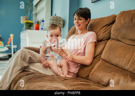 Mitte nach Mutter saß auf einem Sofa im Wohnzimmer. Das baby saß oben auf dem Schoß, nachdem er eine Flasche windet. Das Baby ist ein Blick auf die Kameras Stockfoto