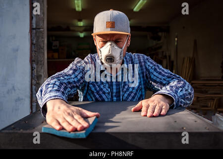 Starke Mann in Arbeitskleidung bemüht und wäscht, Polituren einen Lappen mit einem hölzernen schwarz Tisch in der Werkstatt Stockfoto