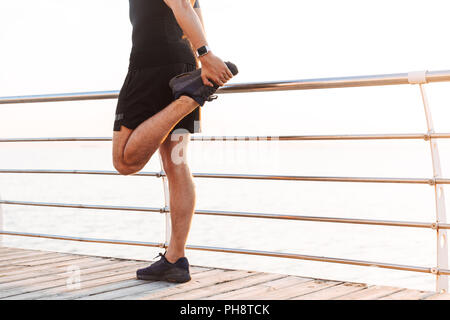 Zugeschnittenes Bild der sportlichen Mann 20 s in Trainingsanzug üben und Strecken die Beine am Pier oder Promenade am Meer am Morgen Stockfoto