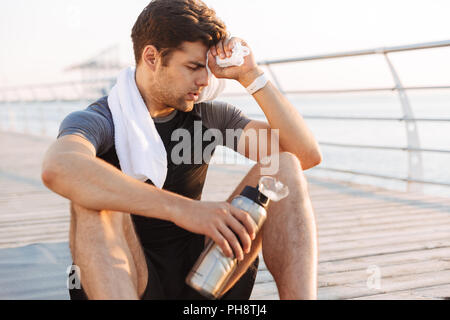 Foto von erwachsenen Sportler 20 s in Trainingsanzug sitzen auf Matten mit Thermoskanne Becher und wischt den Schweiß von der Stirn nach dem Workout auf hölzernen Pier am Meer in Mo Stockfoto