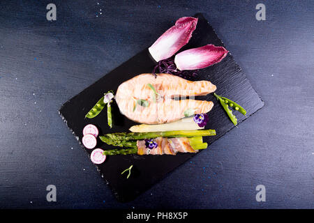 Gegrillter Lachs im Ofen gebacken, mit grünem Spargel im Speckmantel auf schwarzem Hintergrund gewickelt. Overhead. Kopieren Sie Platz. Stockfoto