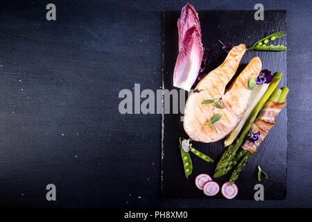 Gegrillter Lachs im Ofen gebacken, mit grünem Spargel im Speckmantel auf schwarzem Hintergrund gewickelt. Overhead. Kopieren Sie Platz. Stockfoto