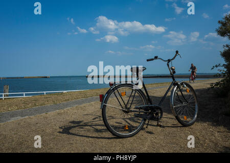 Yachthafen Langeoog bei Niedrigwasser. Deutschland. Traditionelle deutsche Stil Fahrrad vor dem Hafen geparkt. Der alte Hafen Mauern, die im Zweiten Weltkrieg gebaut wurden, WWII, können am Horizont, die auf das Meer gesehen werden. Es ist ein Sonniger Tag casting starke Schatten aus der Fahrrad- und baumhecke mit nur ein Paar weiße geschwollene Wolken im klaren Himmel. Ein Tourist ist Radfahren in die Szene entlang der schmalen Harbour Road. Der Grünstreifen sind ausgetrocknet und ihre Farbe verloren in der Hitzewelle 2018. Das tiefblaue Meer steht in Kontrast zum warmen, blauen Himmel. Stockfoto