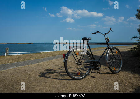 Yachthafen Langeoog bei Niedrigwasser. Deutschland Deutschland. Im traditionellen deutschen Stil Fahrrad neben dem Hafen geparkt. Der alte Hafen Mauern, die im Zweiten Weltkrieg gebaut wurden, WWII, können am Horizont, die auf das Meer gesehen werden. Es ist ein Sonniger Tag casting starke Schatten aus der Fahrrad- und baumhecke mit nur ein Paar weiße geschwollene Wolken im klaren Himmel. Der Grünstreifen sind ausgetrocknet und ihre Farbe verloren im Jahr 2018 Hitzewelle. Das tiefblaue Meer steht in Kontrast zum warmen, blauen Himmel. Stockfoto