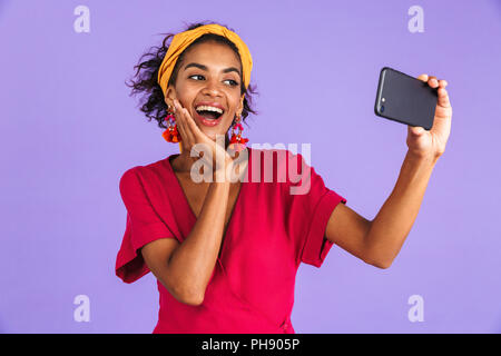 Freudige afrikanische Frau im Kleid, selfie auf dem Smartphone berühren ihre Wange über lila Hintergrund Stockfoto