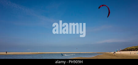 Am Strand Langeoog. Deutschland Deutschland. Eine Person ist Kitesurfen am Wasser entlang auf den Sandstrand. Ein schmaler Streifen des Meeres erstreckt sich entlang der Strand, der Idee für Wassersport. Der kitesurfer Kite erreicht über dem Strand, wo Urlauber sind zu Fuß. Die Sanddünen erweitern aus der rechten Seite des Bildes und die Wind- oder Kitesurf Schule kann unter Flaggen am Horizont am Strand direkt vor den Dünen gesehen werden. Ein einsamer Tourist auf der Meerseite des Strand Uhren die Erfahrene Kitesurfer. An einem sonnigen Tag mit hauptsächlich klarer blauer Himmel exc fotografiert. Stockfoto