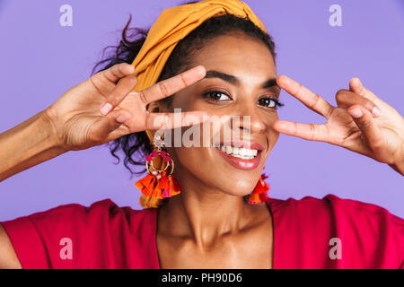 Nahaufnahme, Porträt eines lächelnden jungen afrikanischen Frau in Stirnband über violette Hintergrund, in dem sie zwei Frieden Gesten Stockfoto